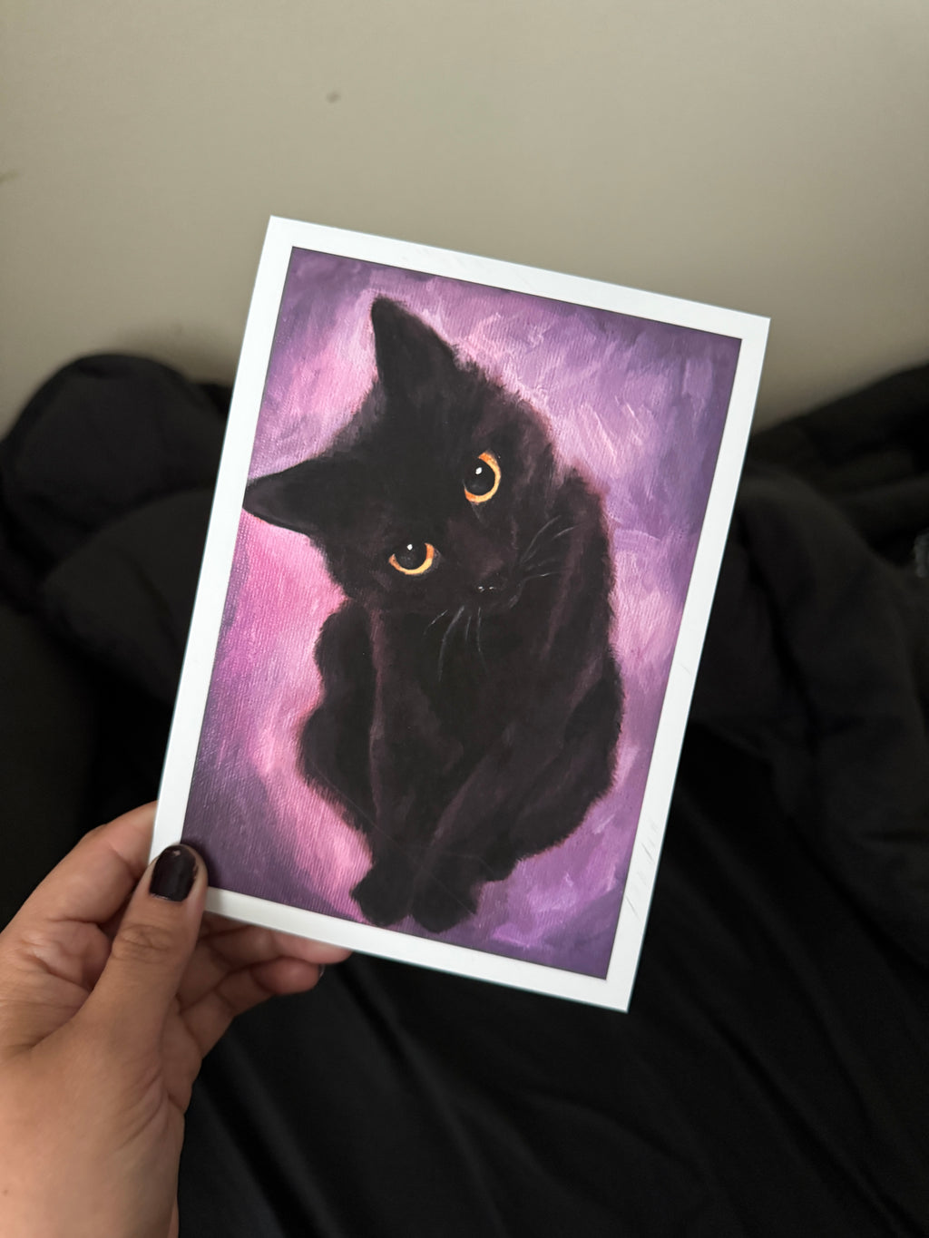 A hand holds a square art print featuring a painted black cat with glowing yellow eyes against a purple and pink background. The print has a white border and is photographed in a casual indoor setting with dark clothing and a beige wall behind it.