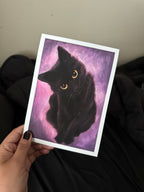 A hand holds a square art print featuring a painted black cat with glowing yellow eyes against a purple and pink background. The print has a white border and is photographed in a casual indoor setting with dark clothing and a beige wall behind it.