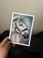  art print featuring a painted portrait of a horse wearing a black bridle against a muted blue-gray background. The print has a white border and is photographed indoors against a beige wall with dark fabric below.