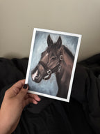 A hand with dark nail polish holds a white-bordered art print featuring a painted brown horse head against a blue-gray background, with dark bedding and a beige wall behind it.