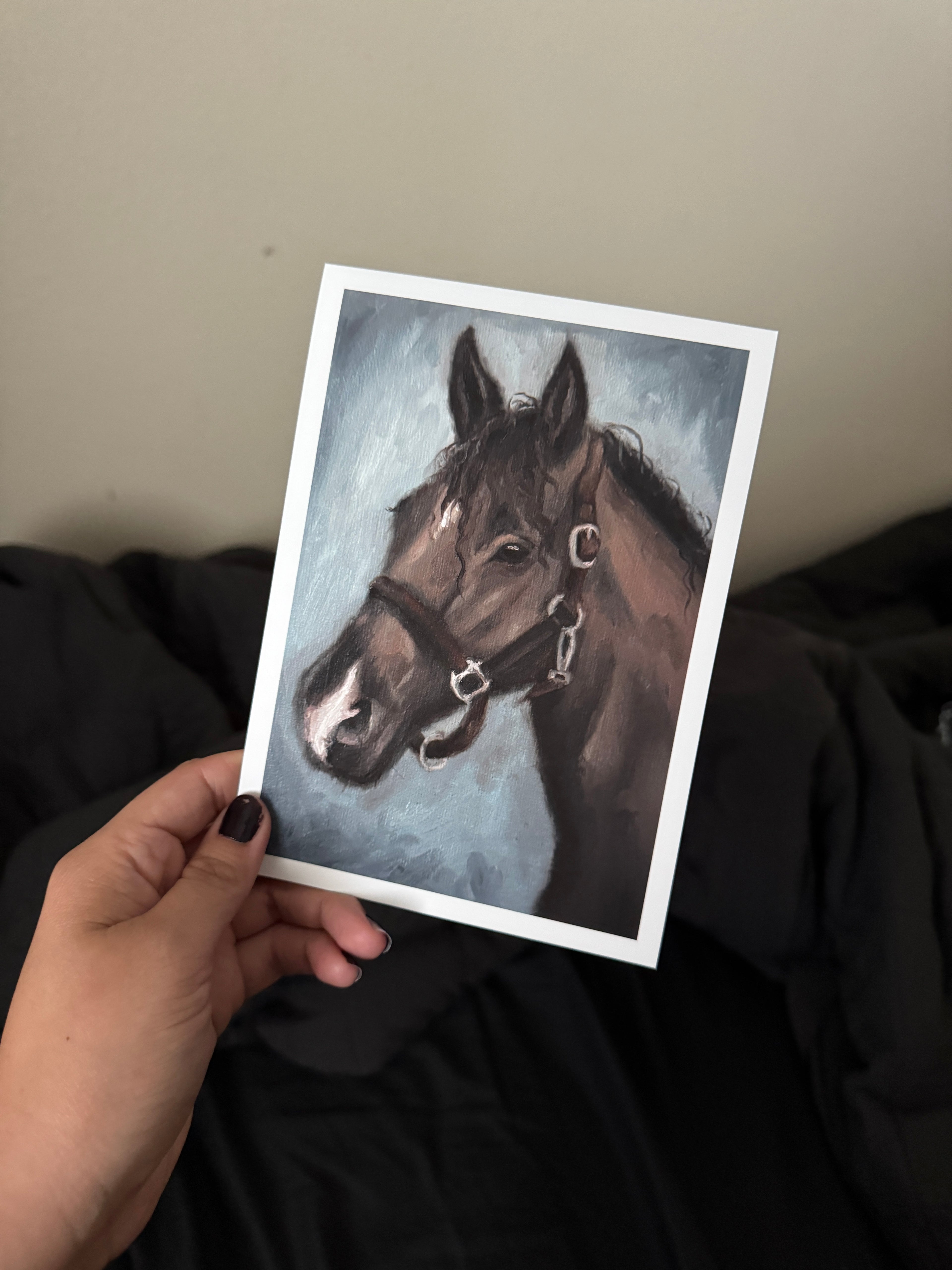 A hand with dark nail polish holds a white-bordered art print featuring a painted brown horse head against a blue-gray background, with dark bedding and a beige wall behind it.