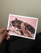 printed art card featuring an oil painting of a deer fawn resting on a pink background, set against a dark couch and beige wall.