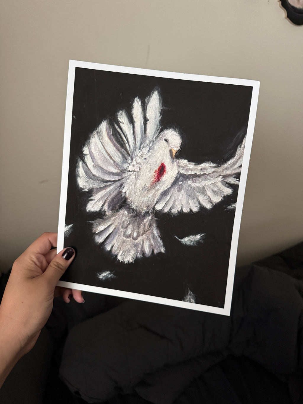 A hand holds a square art print featuring a white dove with outstretched wings on a black background, with a small red accent near the chest. The photo is taken indoors against a beige wall with dark clothing below.
