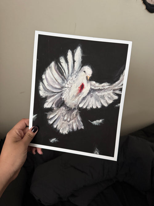 A hand holds a square art print featuring a white dove with outstretched wings on a black background, with a small red accent near the chest. The photo is taken indoors against a beige wall with dark clothing below.