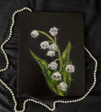 Small painting of lilies of the valley on a dark surface with a pearl necklace.