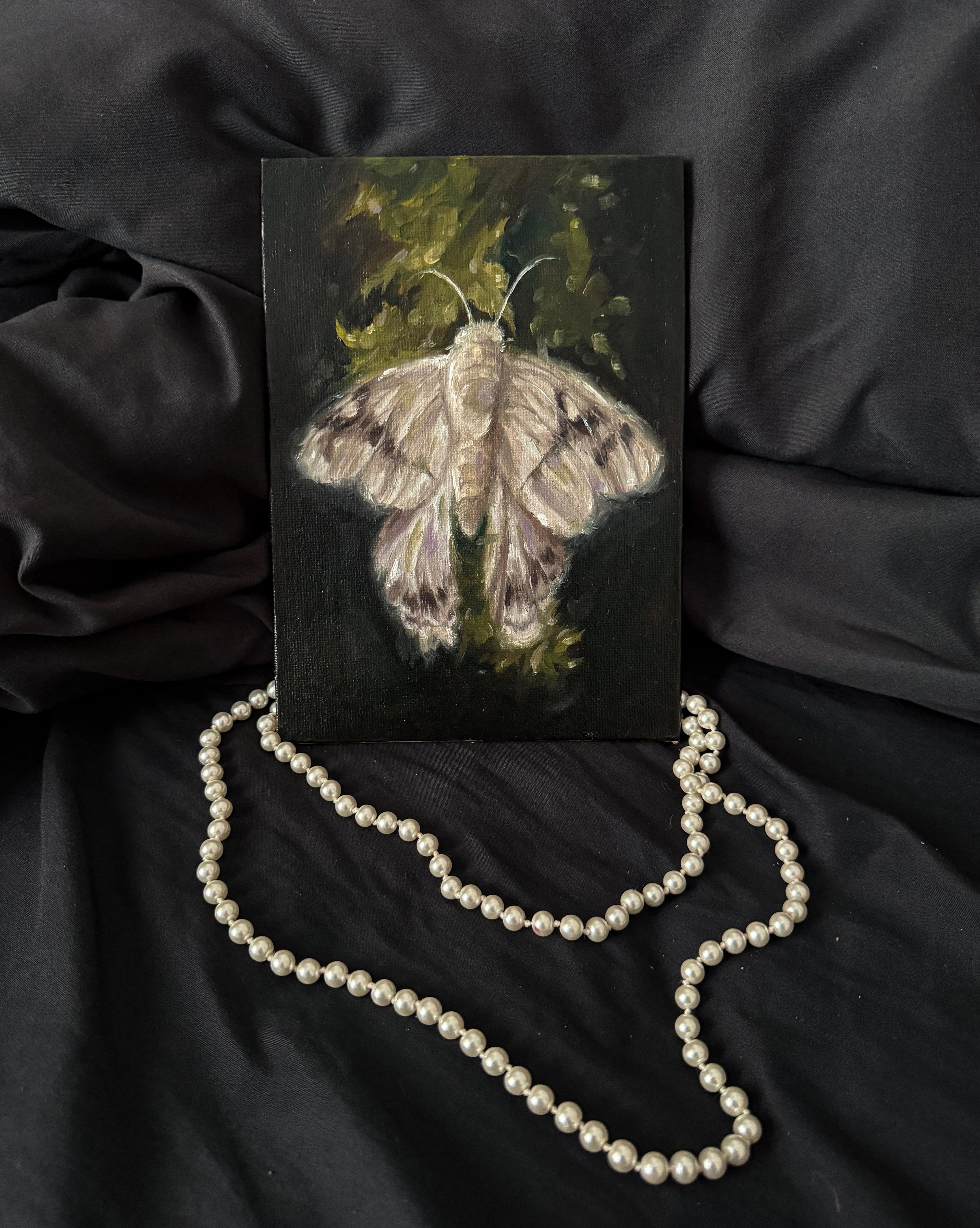 Painting of a moth on a dark surface with a pearl necklace laying besides it